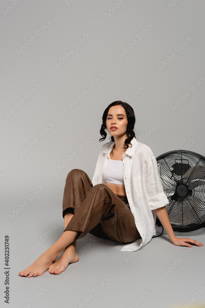 Full length of woman with crossed legs looking at camera while sitting near fan on grey Full length of woman with crossed legs looking at camera while sitting near fan on grey