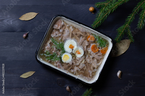 pork jelly with egg, carrot, pea, dill in pot on black wooden background with new year tree brancjes, spices 