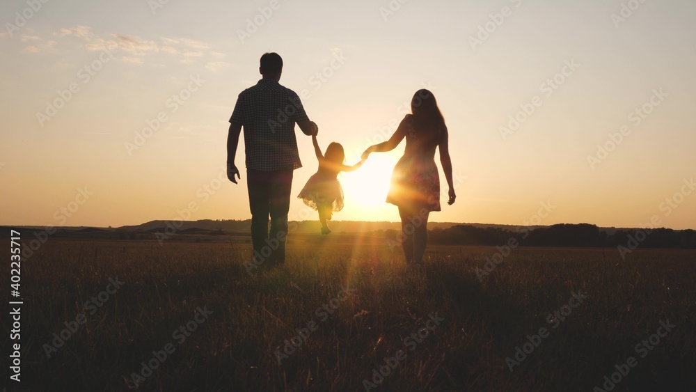 Child plays with dad and mom on field in sunset light. Family and childhood. Little daughter jumping holding hands of dad and mom in park on background of sun. Walking with small kid in nature.