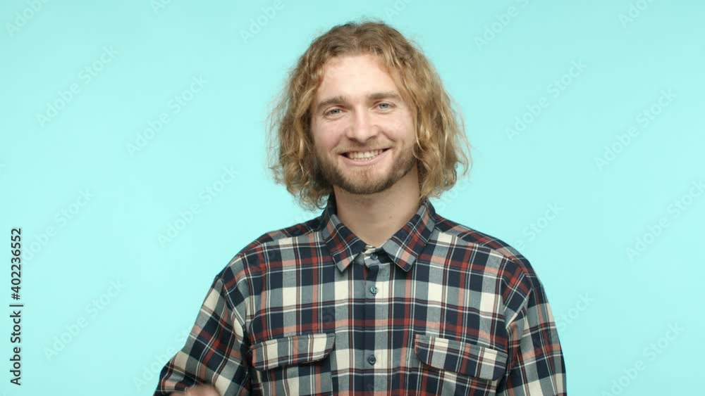 Slow motion of friendly blonde hipster guy saying hello, waving hand and saying hi to camera, smiling cheerful, standing in checked shirt over turquoise background Slow motion of friendly blonde hipster guy saying hello, waving hand and saying hi to camera, smiling cheerful, standing in checked shirt over turquoise background