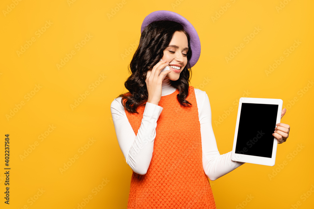 brunette woman in beret talking on cellphone while looking at digital tablet isolated on yellow brunette woman in beret talking on cellphone while looking at digital tablet isolated on yellow