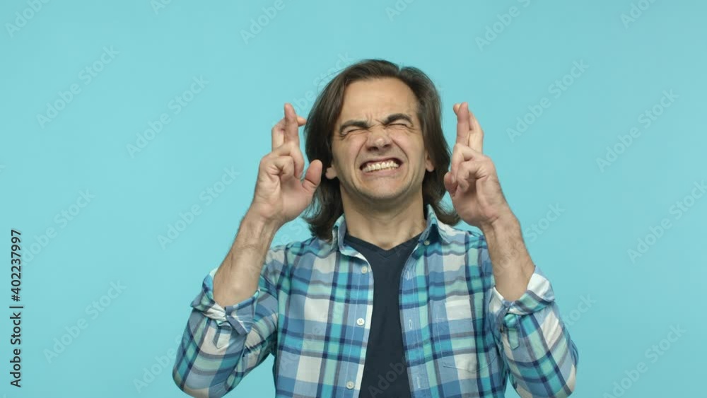 Slow motion of mature 35s man in casual checked shirt, begging and pleading, making wish with crossed fingers, standing hopeful against blue background Slow motion of mature 35s man in casual checked shirt, begging and pleading, making wish with crossed fingers, standing hopeful against blue background