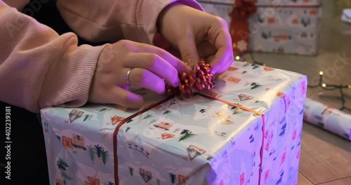 Girl female hand decoration a holiday Christmas gift box with a beautiful funny wrapper and thin red ribbon, tape with a bow in center. Decor paper and light on the background, family happy atmosphere