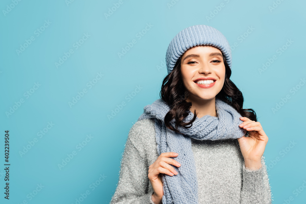  brunette woman in winter outfit touching scarf while looking at camera isolated on blue