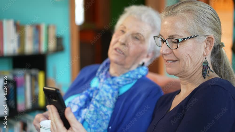 Elderly senior woman and mature daughter drink tea looking at smartphone in living room. Elderly senior woman and mature daughter drink tea looking at smartphone in living room.