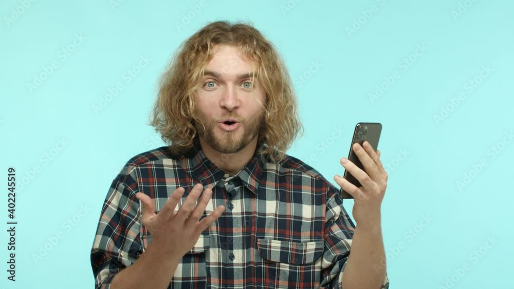 Slow motion of attractive blond guy with beard reading smartphone screen with surprised and happy face, looking amazed and rejoicing, winning online, standing over turquoise background Slow motion of attractive blond guy with beard reading smartphone screen with surprised and happy face, looking amazed and rejoicing, winning online, standing over turquoise background