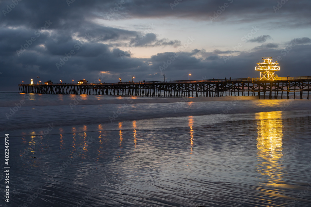 A brightly lit ocean pier on a cold December evening casts reflections on the smooth beach waters below.