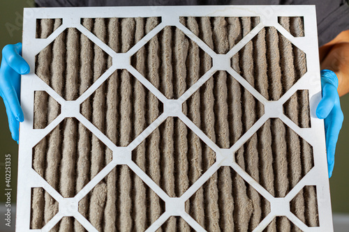 A person wearing blue gloves is holding a heavily clogged dirty air filter in hands before replacing it with the new one. Paper based, card board frame plated filters are used in hvac, ac , furnaces.