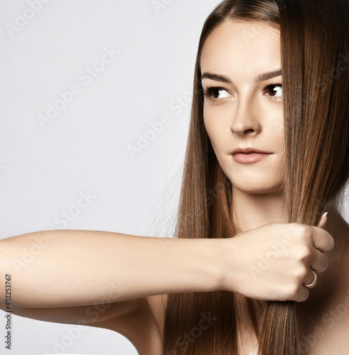 Portrait of sensual young woman holding strand of her long silky hair in her fist demonstrating its strength and health and looking at copy space over grey background. Haircare, beauty, wellness