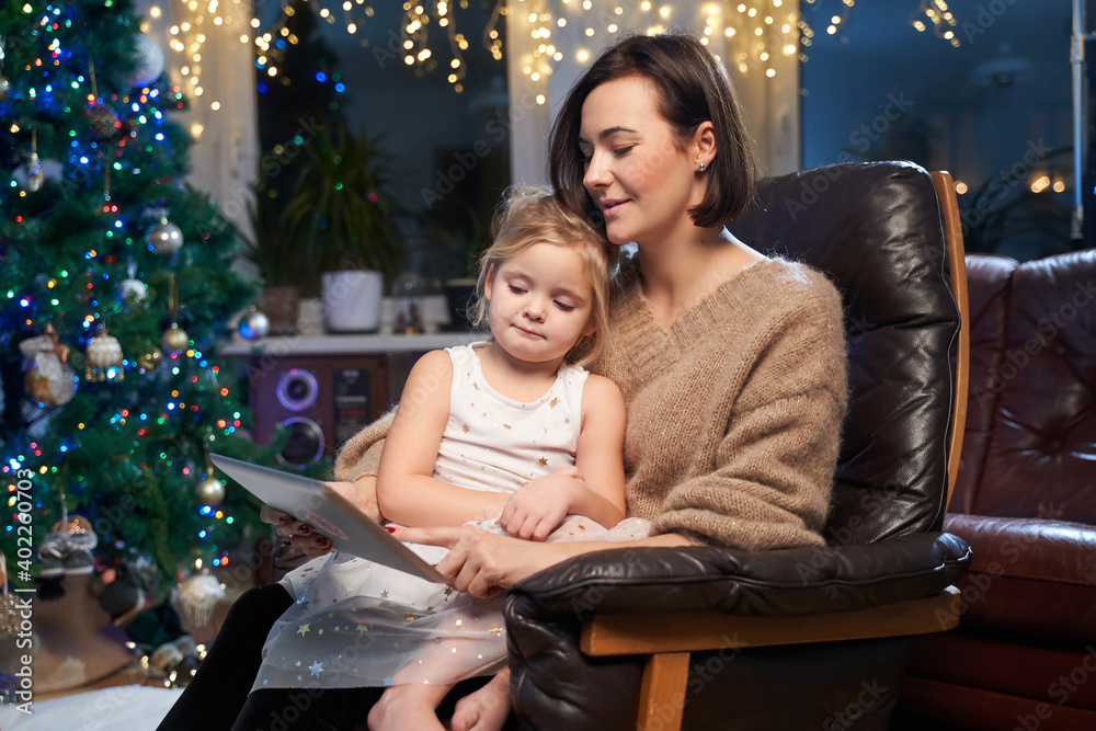 Atmospheric and shiny room with christmas tree and lights and little girl celebrating New Year with her mother sitting on leather armchair with tablet. Atmospheric and shiny room with christmas tree and lights and little girl celebrating New Year with her mother sitting on leather armchair with tablet.