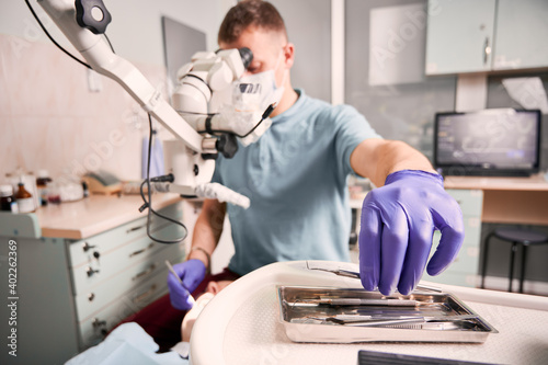 Close up of male dentist hand in sterile glove holding metal dental explorer while checking patient teeth. Stomatologist taking instrument while sitting near microscope. Concept of dentistry.