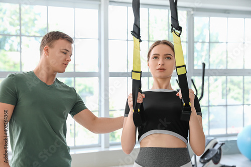 Coach helping young woman to train with TRX straps in gym
