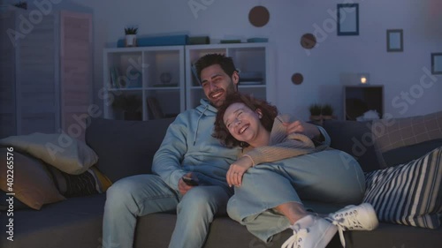 Tracking right of loving diverse couple cuddling on couch at home at night, smiling, cuddling, laughing at comedy on TV. Red-haired Caucasian girl leaning on chest of loving Mixed-Race boyfriend
