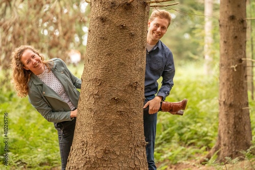 Beautiful young couple playing in the forest, man and woman are half hiding behind a tree