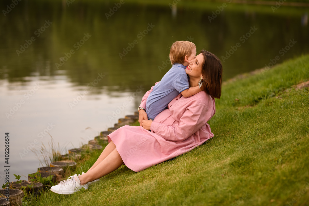 Fototapeta premium Woman with her son on the bank of the pond. They sit huddled. The calm water of the pond reflected scenic shore. Boy and woman admiring the scenery. Peace and harmony