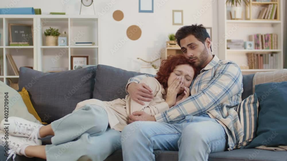 Tracking right shot of Mixed-Race young brown-eyed man cuddling copper-haired Caucasian girlfriend crying over sad movie. Couple sitting on couch in apartment Tracking right shot of Mixed-Race young brown-eyed man cuddling copper-haired Caucasian girlfriend crying over sad movie. Couple sitting on couch in apartment