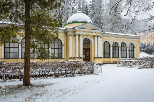 Marianske Lazne, Czech Republic - December 29 2020: Winter view of the Neo-Classical forest spring pavilion with yellow facade standing in a park. Snow covering trees, ground and the building.
