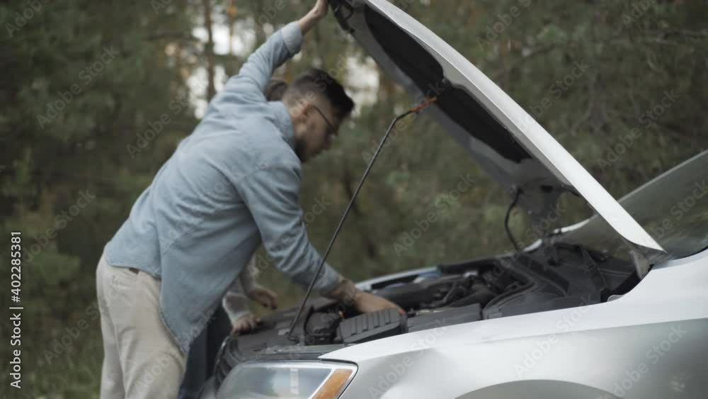 Side view of young Caucasian man in eyeglasses looking at broken car engine in open hood and talking to woman coming. Stressed couple having accident travelling in automobile. Road trouble. Side view of young Caucasian man in eyeglasses looking at broken car engine in open hood and talking to woman coming. Stressed couple having accident travelling in automobile. Road trouble.