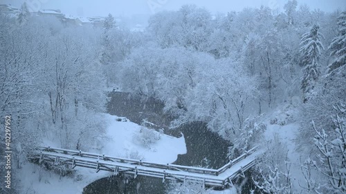 Snowstorm in old medieval town Kranj, Slovenia in winter season. Snow covering Kokra canyon and trees. Small wooden bridge over narrow river. Elevated view. Static shot, real time, wide angle