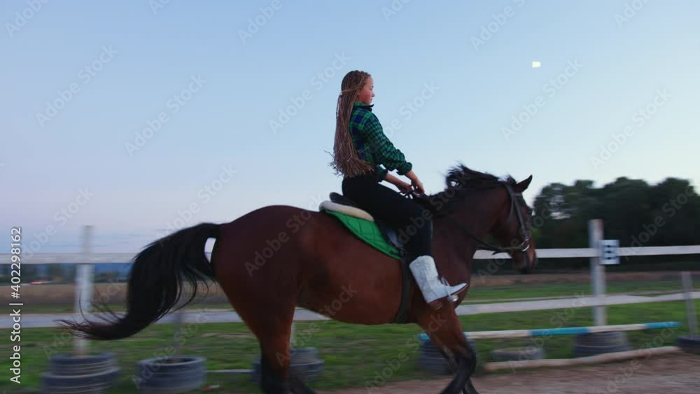 A teenage girl galloping on horseback at outdoor manege. A young girl riding a horse on the farm at dusk A teenage girl galloping on horseback at outdoor manege. A young girl riding a horse on the farm at dusk