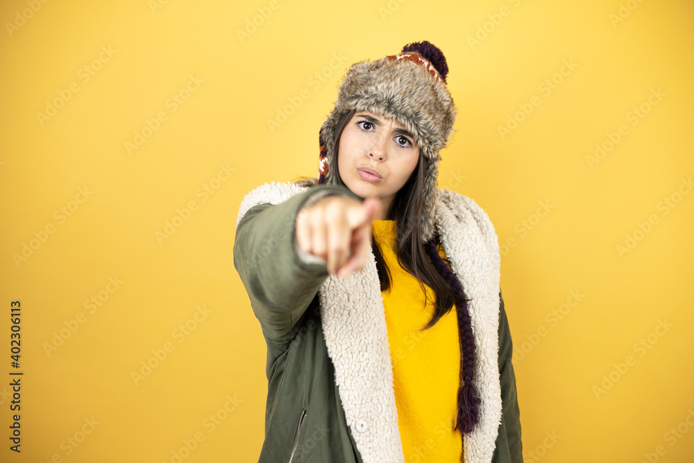 Young beautiful woman wearing a hat and a green winter coat over yellow background pointing to the front with finger Young beautiful woman wearing a hat and a green winter coat over yellow background pointing to the front with finger