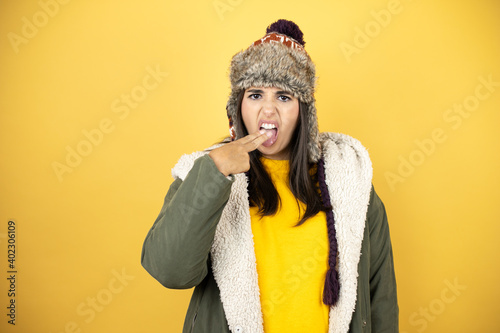 Fotografie Young beautiful woman wearing a hat and a green winter coat over yellow backgrou