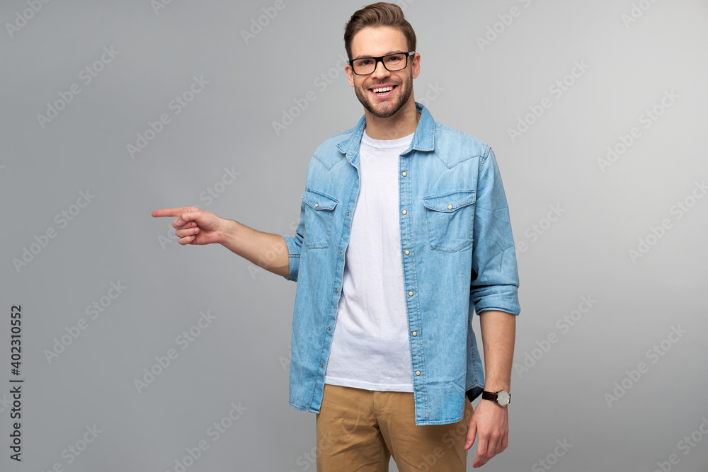 Fototapeta premium Happy young handsome man in jeans shirt pointing away standing against grey background