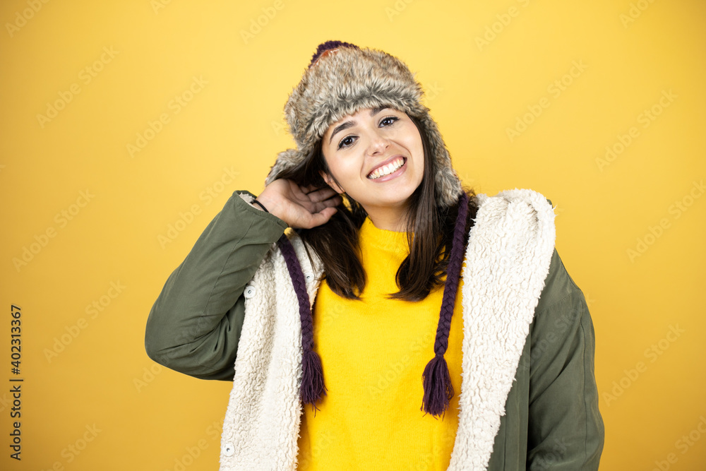 Young beautiful woman wearing a hat and a green winter coat over yellow background smiling to the camera Young beautiful woman wearing a hat and a green winter coat over yellow background smiling to the camera