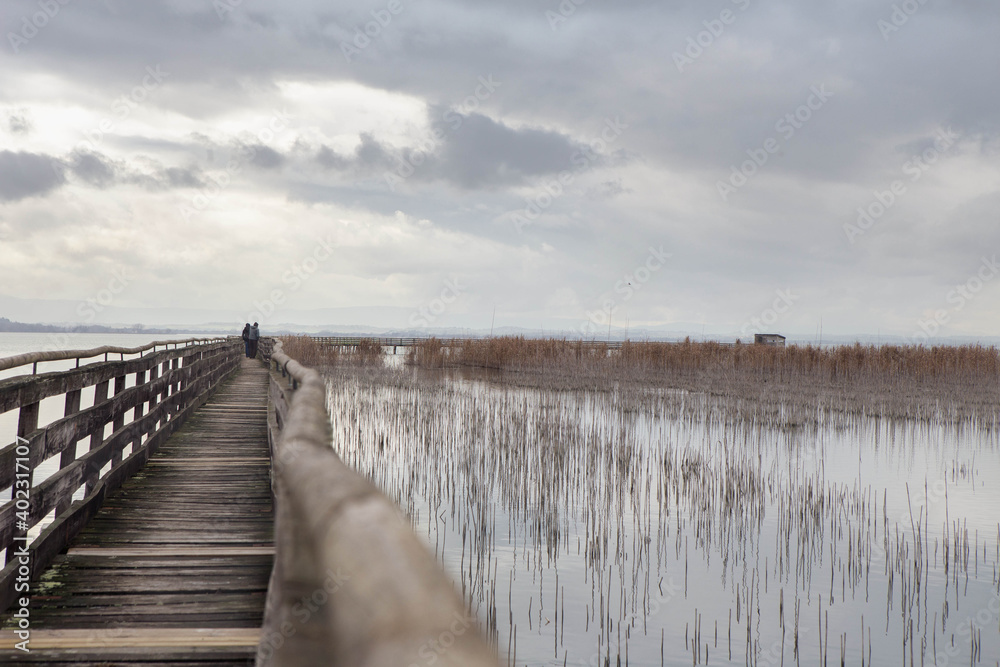Naklejka premium People walking on wooden Pier on Trasimeno lake in a cloudy winter day, in the naturalistic oasis of
