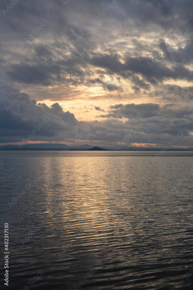 Vetical landscape of Trasimeno lake at sunset  with Isola Polvese in the background,  beautiful cloudy sky reflected in the water, Umbria Italy.