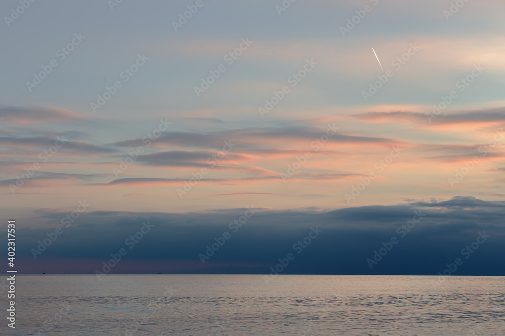 Horizonal sea scape at dusk, with soft pink clouds, blue horizon light and plane trail in the sky.