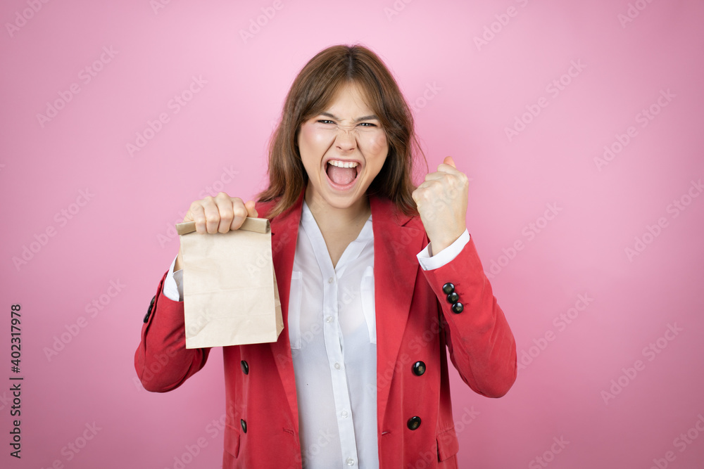 Young business woman over isolated pink background holding a paper bag very happy and excited making winner gesture with raised arms, smiling and screaming for success Young business woman over isolated pink background holding a paper bag very happy and excited making winner gesture with raised arms, smiling and screaming for success