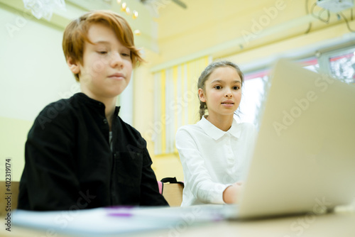 A girl in a white shirt and a boy in a black sweater study online using a computer