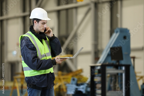 Young man, factory engineer in a work dress and white hard hat holding smartphone and laptop organizing and controlling work process in a industrial factory. 