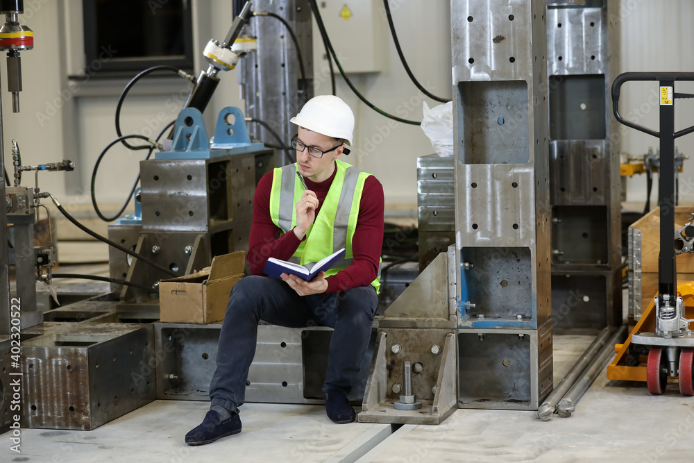 Young man, factory engineer in red pullover and white hard hat holding notebook and pen organizing and controlling work process in a industrial factory. 