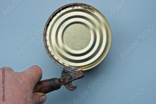 hand holds an old can opener that opens a can with a yellow lid on a gray table photo