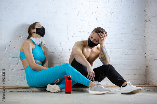 Resting. Professional athletes training on brick wall background wearing face masks. Sport during quarantine of coronavirus worldwide pandemic. Young couple practicing in gym safe using equipment.