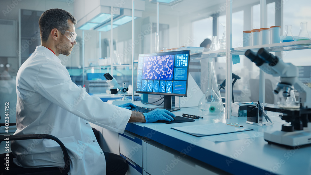 Medical Science Laboratory: Microbiologist Working on Computer with Display Showing Gene Editing Interface. Diverse Multi-Ethnic Team of Biotechnology Scientists Developing Drugs. Medical Science Laboratory: Microbiologist Working on Computer with Display Showing Gene Editing Interface. Diverse Multi-Ethnic Team of Biotechnology Scientists Developing Drugs.