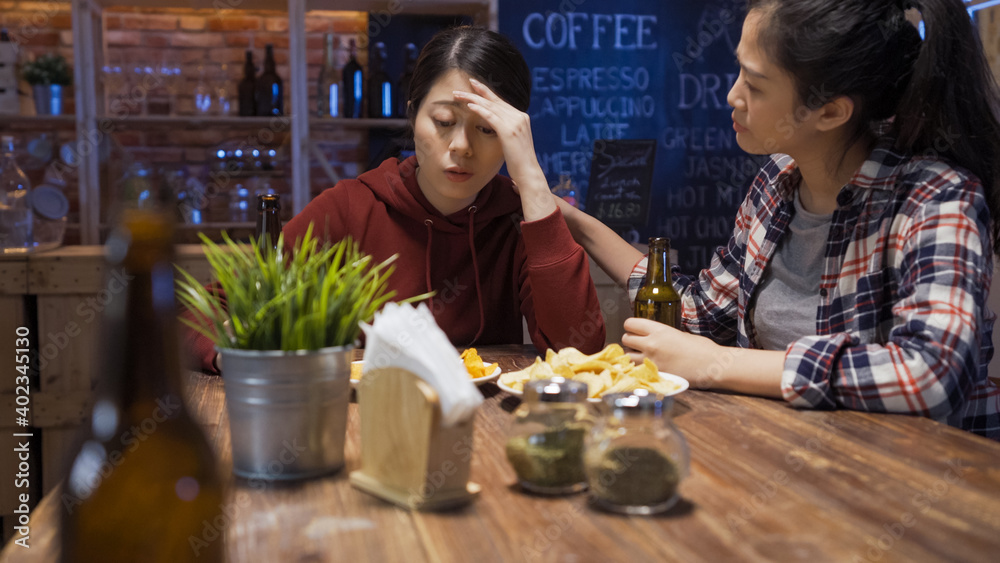 elegant woman comforting depressed friend in bar at night. people friendship consoling alcoholism problem concept. two young upset girl friends in stressed emotion drinking alcohol and beer in pub.