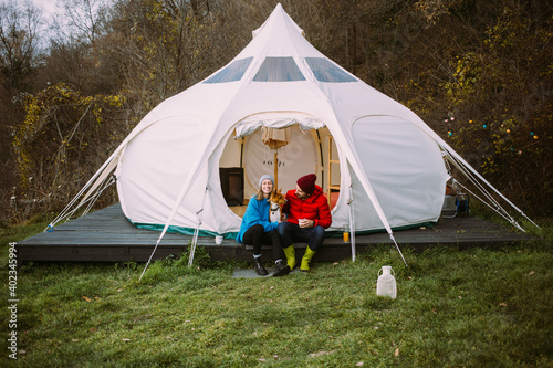 Couple of vacation goers campers sit in front of big glamping tent. Happy couple on romantic weekend getaway with pet dog. Fun and luxury camping experience in nature