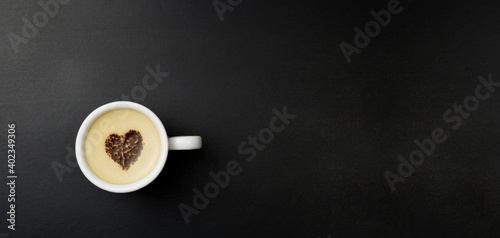 Cup of cappuccino in the center of the heart, on a black background with copy space. Chocolate heart in a cup with a hot coffee drink.