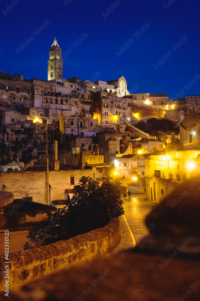 Night View of a street of the ancient stone city of  Matera at dusk. In the background the Sasso Baritano with the tower of Matera Cathedral Maria Santissima della Bruna illuminated. Basilicata, Italy