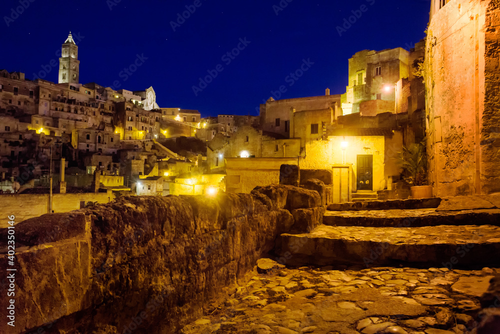 Night view of a street of the ancient stone city of  Matera, In the background the Sasso Baritano with the tower of Matera Cathedral Maria Santissima della Bruna illuminated. Basilicata, Italy, Europe
