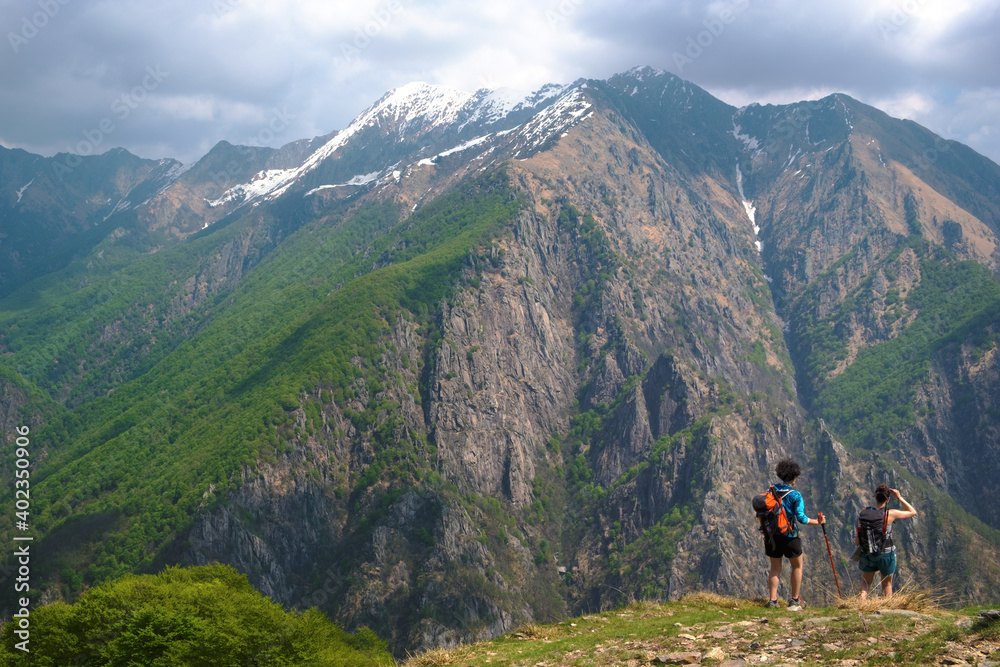 Couple of young women hiking along a mountain path, in the background the wild panorama of  the Val Grande National Park, Europe, Piedmont Italy.