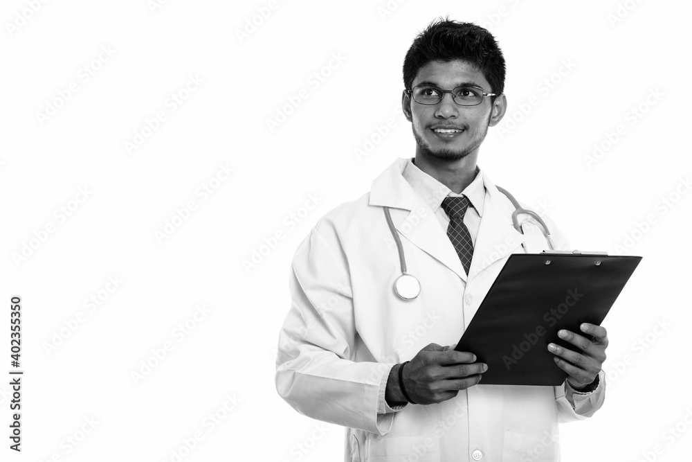 Studio shot of young happy Indian man doctor smiling and holding clipboard while thinking Studio shot of young happy Indian man doctor smiling and holding clipboard while thinking