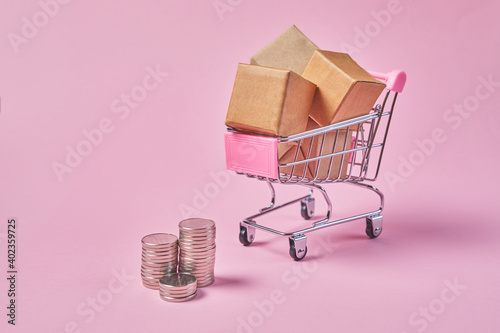 Shopping cart with package boxes and coins on a pink background. Shop trolley full of parcels.