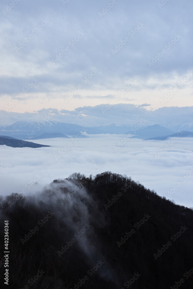 Low clouds, view of winter forest and mountains from observation deck, vertical picture of amazing natural phenomenon. Beautiful unreal panoramic view of mountains in clouds.