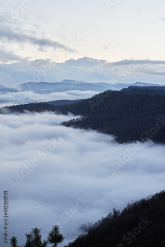 Low clouds, view of winter forest and mountains from observation deck, vertical picture of amazing natural phenomenon. Beautiful unreal panoramic view of mountains in clouds.