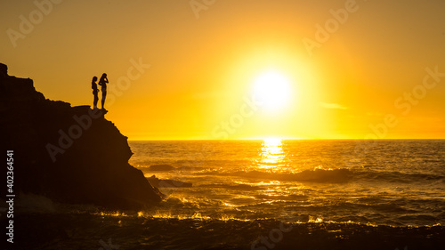 Two Girl Silhouettes On A Cliff With Orange Sky Sunset