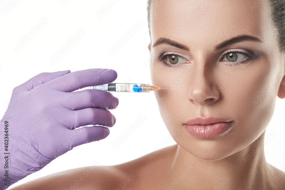 Beautician's hand in purple glove holding a syringe near beautiful woman's face. Portrait isolated on a white background. Anti-aging treatment.Injection cosmetology Beautician's hand in purple glove holding a syringe near beautiful woman's face. Portrait isolated on a white background. Anti-aging treatment.Injection cosmetology